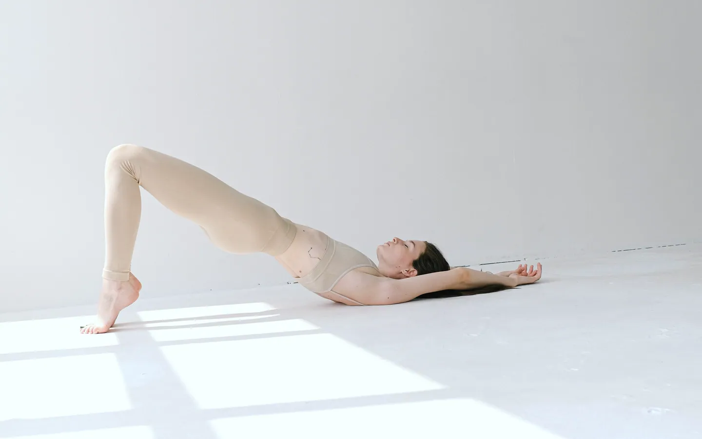 A woman lies on a white floor and lifts her hips into a bridge, with sunlight falling across the room.