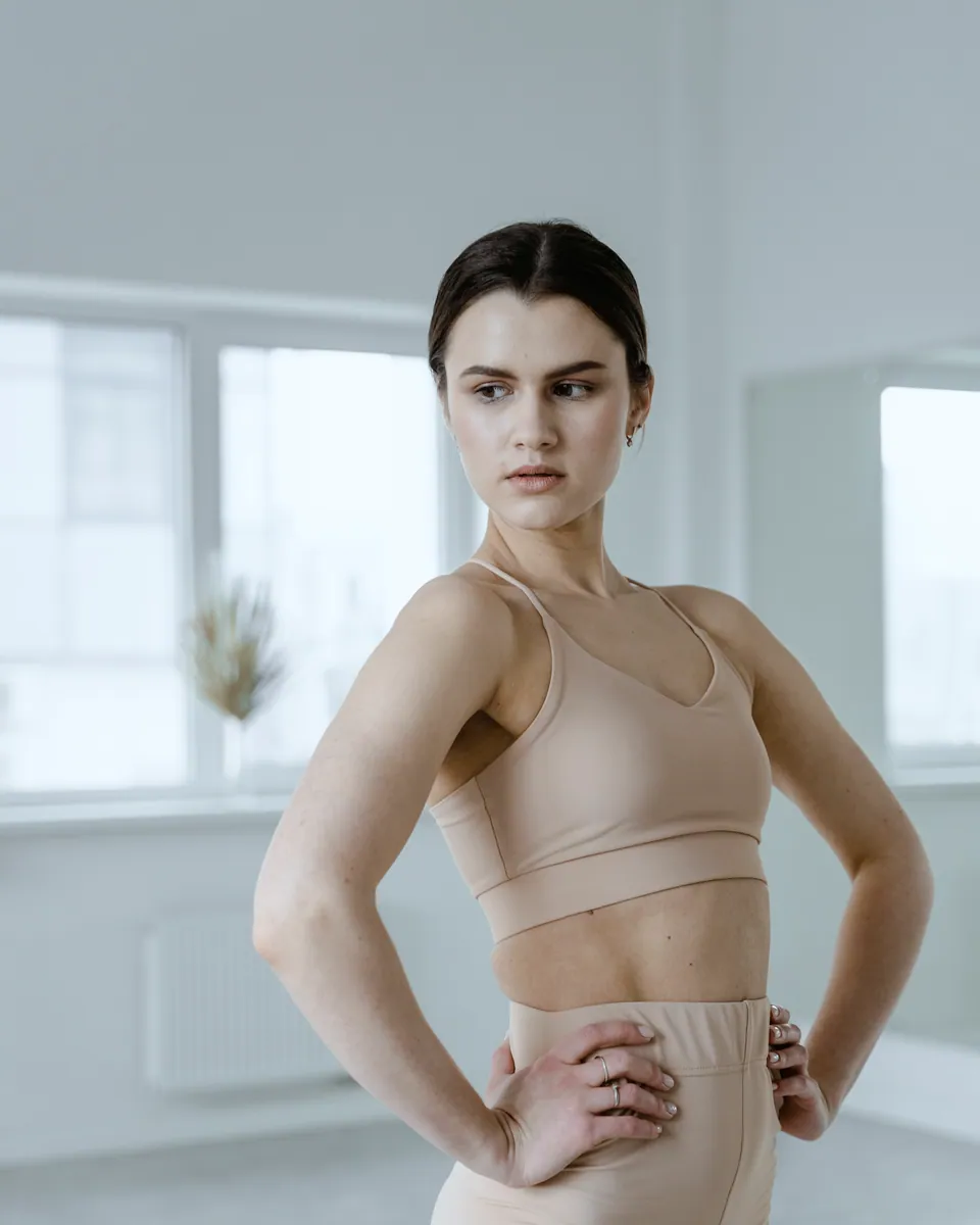 A woman in a beige sports top and leggings stands with her hands on her hips in a bright, empty studio.