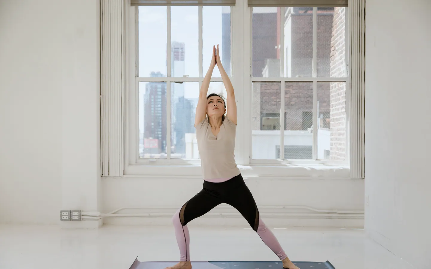 A woman stands on a mat with her arms raised above her head and her palms together, in front of tall white-framed windows.