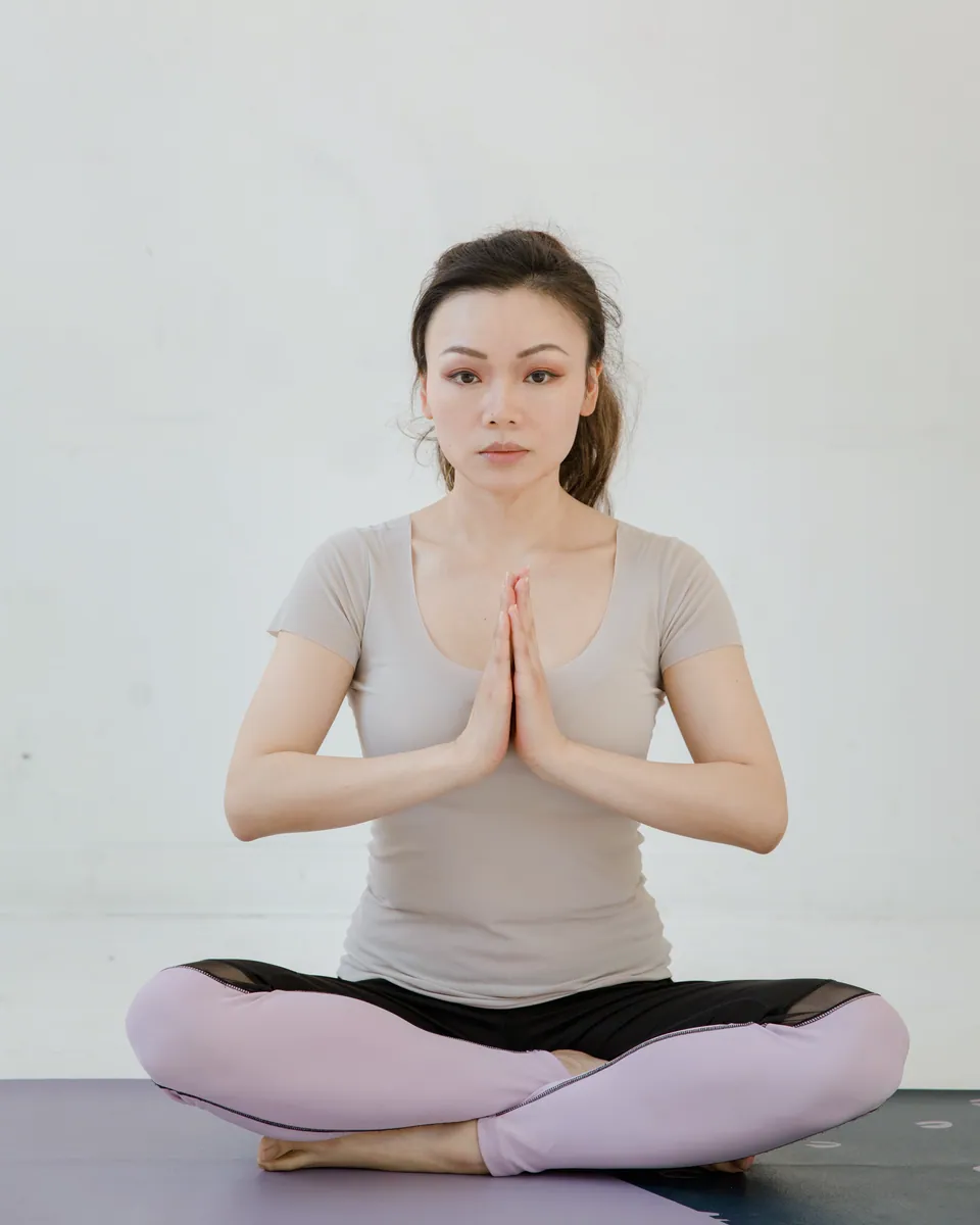 A woman sits cross-legged on a mat with her palms pressed together and her eyes closed, against a plain white wall.