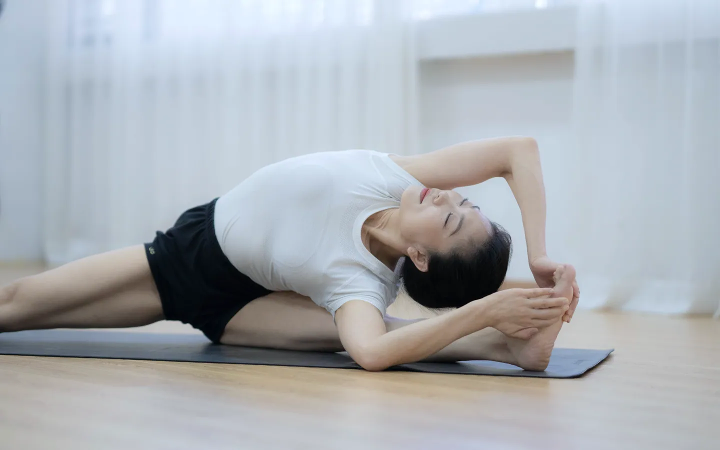 A woman in a white top sits on a dark mat and bends sideways over one outstretched leg, with sheer curtains and soft daylight behind her.